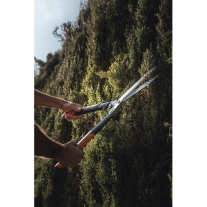 Taille-haie à crémaillère Fiskars HSX92 taillant une haie. Coupe-haie manuel avec poignées grises et lames argentées.