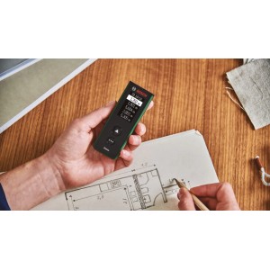 Hand hält grünen Bosch Zamo Laser-Entfernungsmesser, zeigt Messwerte. Person plant mit Bauplan, nutzt präzises Messwerkzeug für Längen.