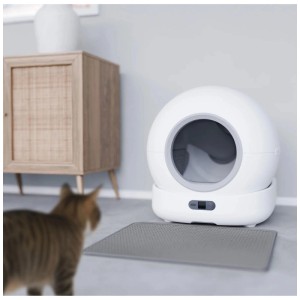 Weisse Hombli Smart-Katzentoilette mit Kamera, kugelförmig, graue Matte davor. Eine Katze blickt auf das moderne Katzenklo.