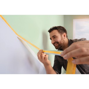 Un homme applique du ruban de masquage jaune tesa® Malerband Perfect+ sur un mur pour des bords de peinture nets.