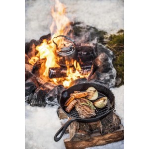 Padella grill Petromax in ghisa con bistecca, verdure e fuoco da campo sullo sfondo.