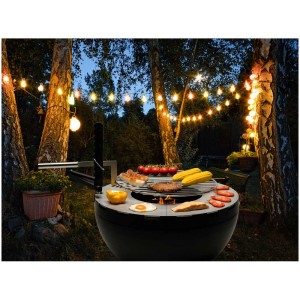Brasero-grill Nouvel Firebowl 80 avec aliments dans le jardin sous guirlande lumineuse.