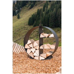Range-bûches rond noir Hestia (Ø 78 cm) en acier, rempli de bois, sur fond de montagne.