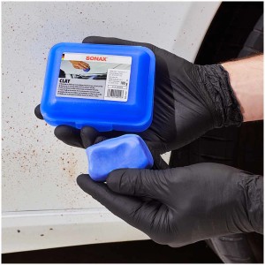 Sonax Clay Reinigungsknete 100g in blauer Dose, gehalten von Händen mit schwarzen Handschuhen.