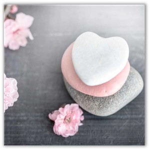 Quadro in vetro Heart Stone: Tre pietre a forma di cuore (bianca, rosa, grigia) con fiori rosa su sfondo grigio.