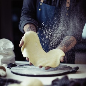 Hände dehnen Pizzateig über einem runden, grauen Keramik-Pizzastein für knusprige Pizza vom Grill oder Backofen.
