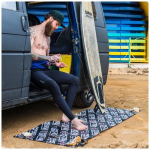 Muc-Off Bike Mat Werkstattmatte 70x209cm: Surfer cremt sich auf der Matte am Strand ein.