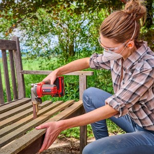 Donna che leviga una panca di legno con la levigatrice a nastro a batteria rossa Einhell TE-BF 18 V Li-Solo. Utensile a batteria con nastro abrasivo.