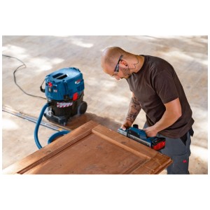 Uomo che pialla una porta con pialla a batteria Bosch Professional GHO 18V-26. Aspirazione polvere con aspirapolvere.