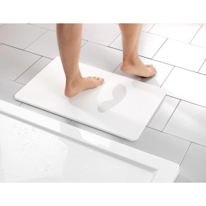 Tappetino da bagno bianco Wenko Simi, 60x39 cm, in diatomite con impronte di piedi, davanti alla vasca da bagno su piastrelle.
