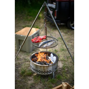 Grille suspendue Petromax sur trépied au-dessus d'un foyer avec poisson et poivrons. Ustensile de grill pour feu de camp.