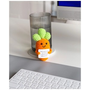 Gehäkelter Glücksbringer Karotte, orange mit grünem Laub, lächelt, hält Schild auf Schreibtisch.