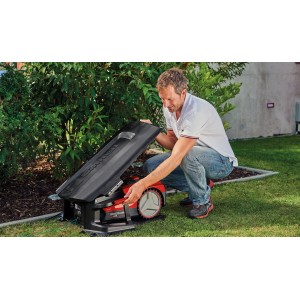 Robot tondeuse Einhell avec garage ouvert dans le jardin, manipulé par un homme.