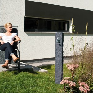 Colonna d'acqua Garantia Granit Darkgranite con rubinetto cromato in giardino, donna seduta accanto.