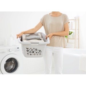 Donna che trasporta cesto biancheria Rotho Flowers Cool Grey con bucato davanti alla lavatrice.