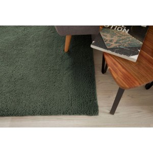 Tappeto a pelo lungo Elvas verde scuro su pavimento in legno chiaro, con poltrona e tavolino. Ambiente accogliente.