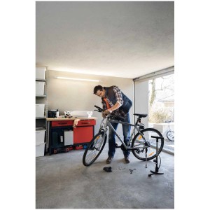 Homme réparant un vélo sous un tube LED Osram dans un garage. Établi et outils visibles.