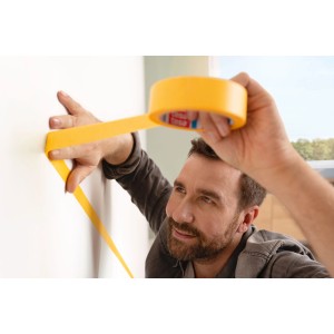 Homme appliquant du ruban de masquage jaune Tesa Malerband Perfect+ sur un mur blanc. Ruban de masquage pour des bords de peinture précis.