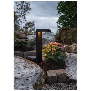 Schwarze LED Solar Pollerleuchte Vicini von Star Trading im Garten zwischen Pflanzen und Steinen.