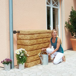Garantia Serbatoio da parete Rocky 400 l Sandstone con donna e annaffiatoio davanti al muro di casa. Barile per la pioggia effetto pietra.