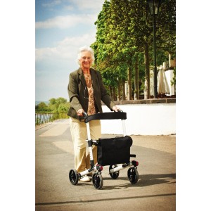 Femme utilisant un rollator Ridder Premium 4 roues à l'extérieur. Rollator argenté avec sac noir.
