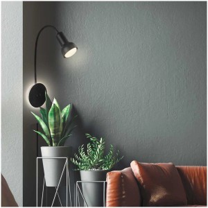 Applique murale LED Briloner noire avec rétroéclairage et spot, montée sur un mur gris au-dessus de plantes et d'un canapé.