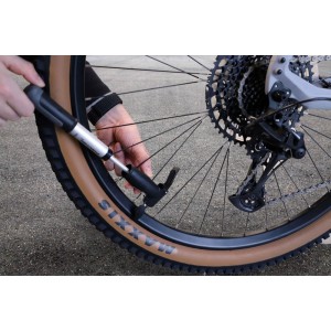 Mini pompa d'aria Fischer Profiplus in azione: Mani gonfiano uno pneumatico da bicicletta. Pompa argento sul copertone marrone.