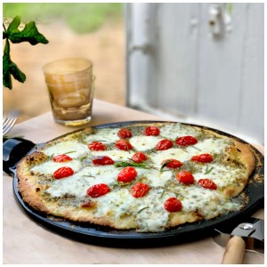 Schwarzer Emile Henry Pizzastein aus Keramik (Ø 37 cm) mit Pizza, Tomaten und Kräutern.