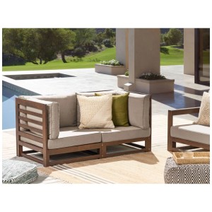 Divano da giardino modulare Beliani Trani in legno di acacia scuro, cuscini chiari, su patio.