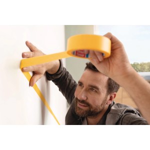 Homme appliquant du ruban de masquage jaune tesa® Malerband Perfect+ sur un mur blanc pour des bords de peinture précis.