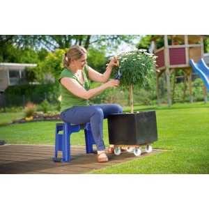 Femme assise sur tabouret de jardin LUX bleu avec coussin de genoux, taillant une plante dans le jardin.
