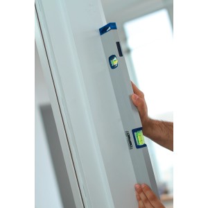 Niveau à bulle LUX Comfort 40 cm en utilisation : une main tient le niveau argenté contre un cadre de porte blanc.