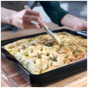 Schwarze Emile Henry Focaccia-Backform aus Keramik mit Focaccia und Rosmarin wird mit Öl bepinselt.